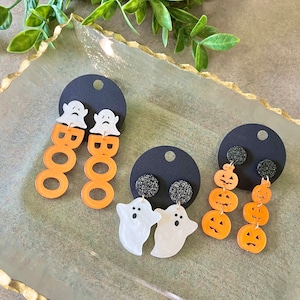 May include: Three pairs of Halloween-themed earrings. The first pair features white ghost charms with the word "BOO" in orange letters. The second pair features white ghost charms with black glitter accents. The third pair features orange pumpkin charms.