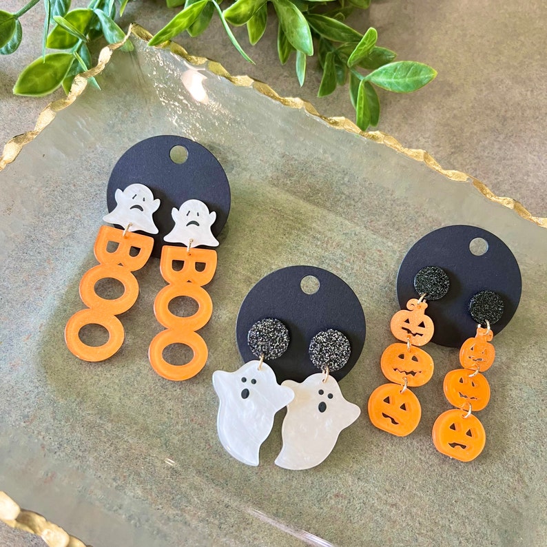 May include: Three pairs of Halloween-themed earrings. The first pair features white ghost charms with the word "BOO" in orange letters. The second pair features white ghost charms with black glitter accents. The third pair features orange pumpkin charms.
