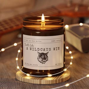 May include: A lit soy wax candle in an amber glass jar with a white label featuring the text 'Smells Like A WILDCATS WIN' and a graphic of a wildcat head. The label also includes 'C'est la Vie Gifts', '100% natural', '9oz', and '50 hours'.