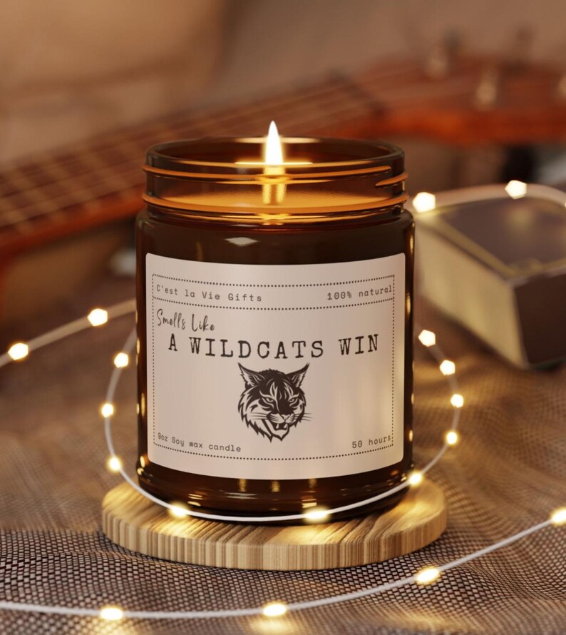 May include: A lit soy wax candle in an amber glass jar with a white label featuring the text 'Smells Like A WILDCATS WIN' and a graphic of a wildcat head. The label also includes 'C'est la Vie Gifts', '100% natural', '9oz', and '50 hours'.