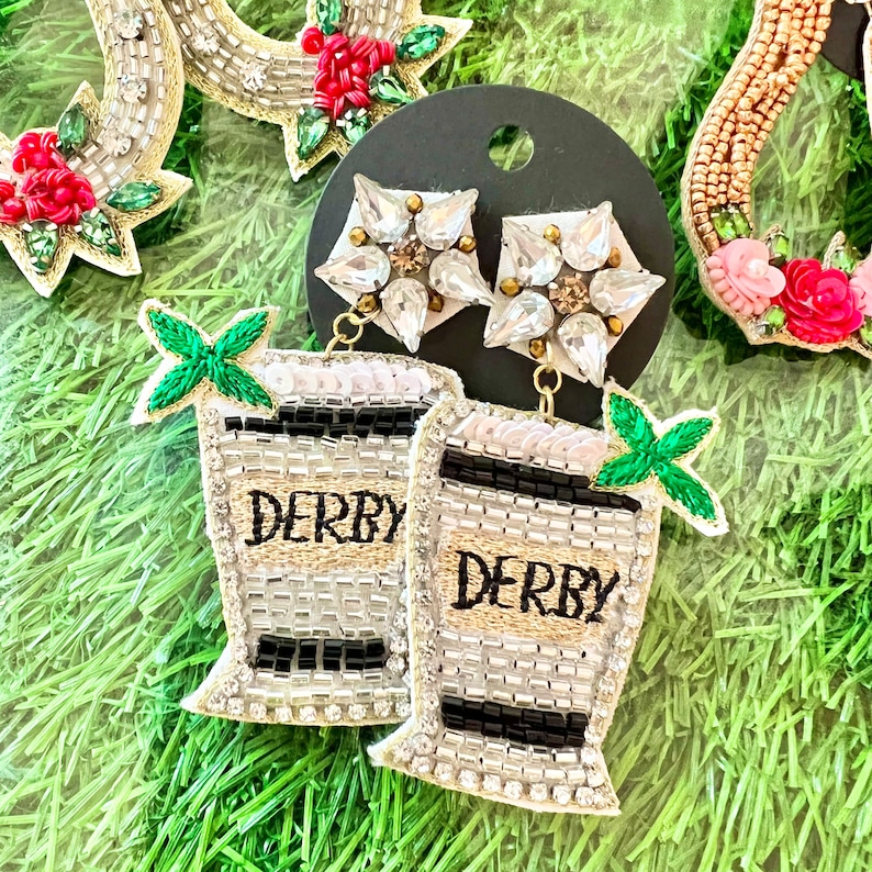 May include: A pair of silver and black beaded earrings with a green leaf charm and the word "Derby" on each earring. The earrings are hanging from a black card.