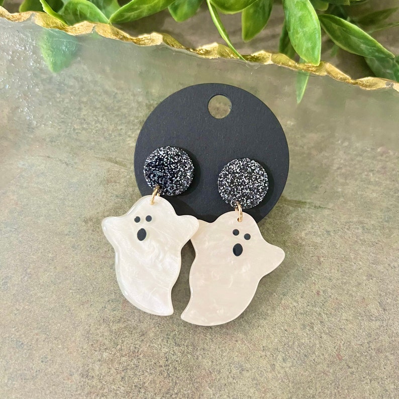 May include: A pair of white ghost-shaped earrings with black eyes and a black glitter accent. The earrings are hanging from a black card.