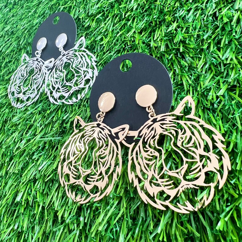 May include: Gold tone tiger head earrings with a cutout design. The earrings have a gold tone post and a clear stone.