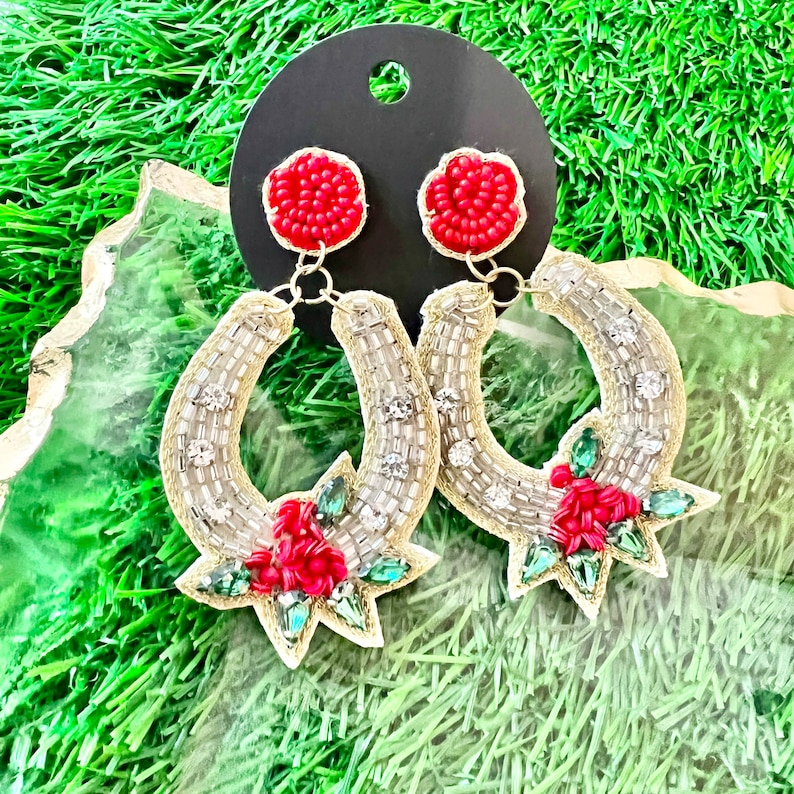 May include: A pair of silver horseshoe-shaped earrings with red and green beaded flowers. The earrings are hanging from a black display card.