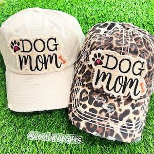 May include: Two distressed baseball caps with the text "Dog Mom" in black lettering on a white patch. One cap is beige and the other is leopard print.