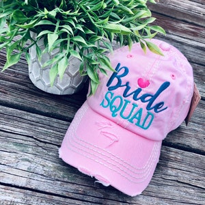 May include: Pink distressed baseball cap with the text "Bride Squad" embroidered in blue and teal with a pink heart.