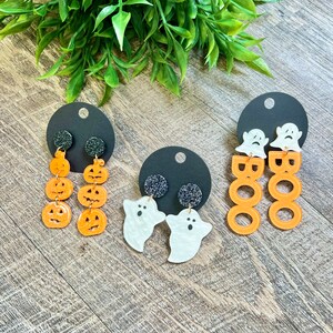 May include: Three pairs of Halloween-themed earrings. One pair features orange pumpkin earrings, another pair features white ghost earrings, and the last pair features orange "BOO" earrings.