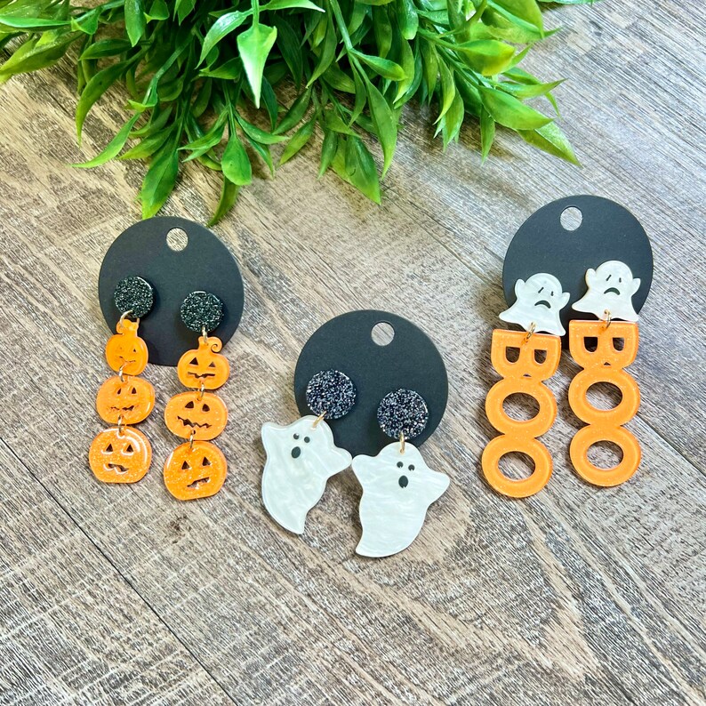 May include: Three pairs of Halloween-themed earrings. One pair features orange pumpkin earrings, another pair features white ghost earrings, and the last pair features orange "BOO" earrings.