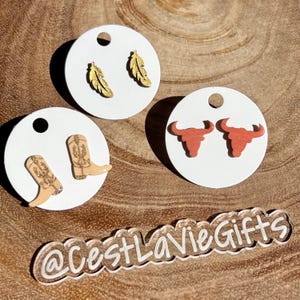 May include: Three sets of earrings on white circular cards. The first set features gold feather earrings. The second set features brown cowboy boot earrings. The third set features red bull head earrings. The cards are on a wooden surface with the text "@CestLaVieGifts" printed on a clear acrylic surface.
