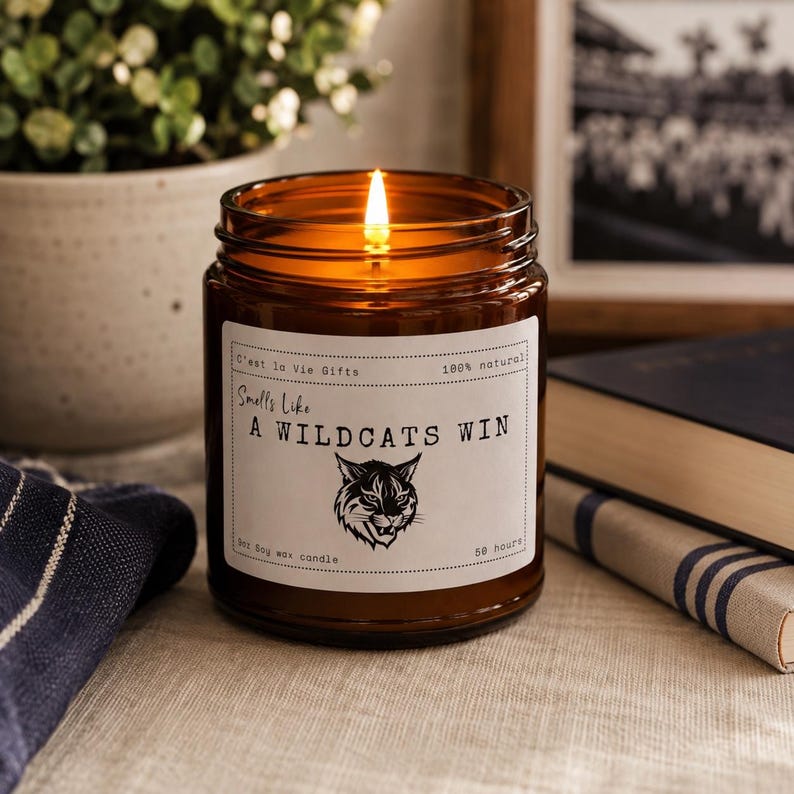 May include: A lit candle in an amber glass jar with a white label featuring the text 'Smells Like A WILDCATS WIN' and a black graphic of a wildcat head. The label also includes the text 'C'est la Vie Gifts', '100% natural', '9oz Soy wax candle'.