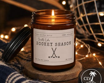 May include: A brown glass jar candle with a black lid and a white label featuring the text Smells Like HOCKEY SEASON, an illustration of two crossed hockey sticks, and the phrases C'est la Vie Gifts, 100% natural, 9oz Soy wax candle, 50 hours.