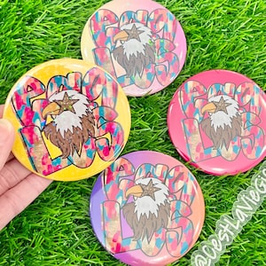 Puede incluir: Cuatro botones redondos con un signo de paz y un diseño de águila. Los botones son amarillos, rosas, morados y rojos con un signo de paz blanco y un águila marrón. Los botones tienen un acabado brillante e iridiscente.