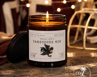 May include: A lit candle in an amber glass jar with a black lid. The white label features the text 'C'est la Vie Gifts', '100% natural', 'SMELLS LIKE A GAMECOCKS WIN', 'For best results, light before kickoff', and an illustration of a fighting rooster.