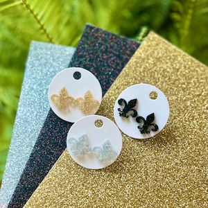 May include: Three pairs of earrings featuring a fleur-de-lis design. The earrings are on white cards with a hole at the top. The earrings are gold, silver, and black.