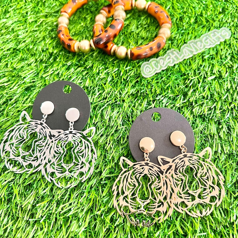 May include: A pair of gold tiger head earrings with a black background. The earrings are on a black card with a gold stud.