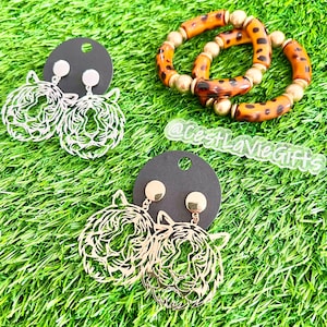 May include: A pair of gold tiger head earrings and a pair of silver tiger head earrings on a black card. The earrings are on a green grassy background. A brown and gold leopard print bracelet is also in the image. The text "@CestLaVieGifts" is visible in the image.
