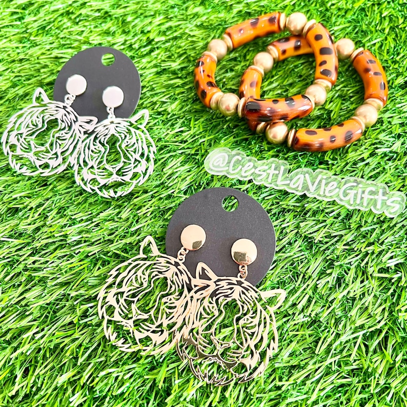 May include: A pair of gold tiger head earrings and a pair of silver tiger head earrings on a black card. The earrings are on a green grassy background. A brown and gold leopard print bracelet is also in the image. The text "@CestLaVieGifts" is visible in the image.