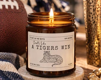 May include: A lit soy wax candle in an amber glass jar with a white label featuring a tiger illustration and the text C'est La Vie Gifts, Smells Like A TIGERS WIN, 100% Natural, 9oz Soy Wax Candle, and 50 Hours printed on the front surface.