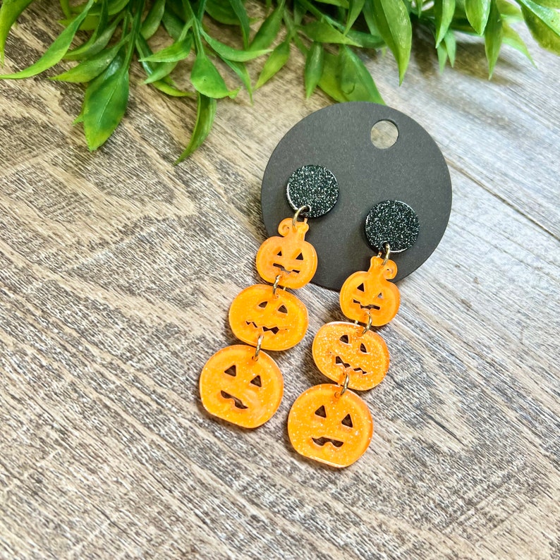 May include: A pair of dangle earrings featuring three stacked orange pumpkin charms with black glitter accents. The earrings are attached to black glitter posts.