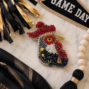 May include: A handcrafted rooster head brooch featuring intricate beadwork and sequins. The design incorporates vibrant red, black, yellow, and white beads with metallic gold accents, creating a textured, multi-dimensional appearance.