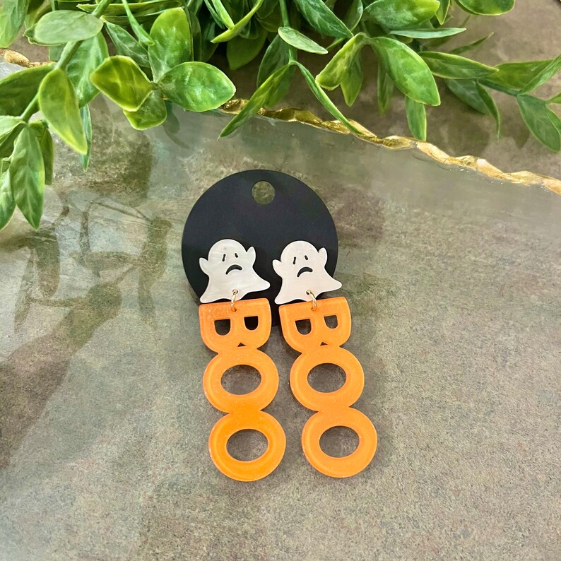 May include: Orange acrylic earrings shaped like the word "BOO" with a white ghost charm on top of each earring.