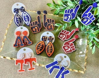 May include: Assortment of beaded earrings featuring various sports-themed designs, including footballs, hands making the number one gesture, and letters. The earrings are in shades of blue, red, orange, and white.