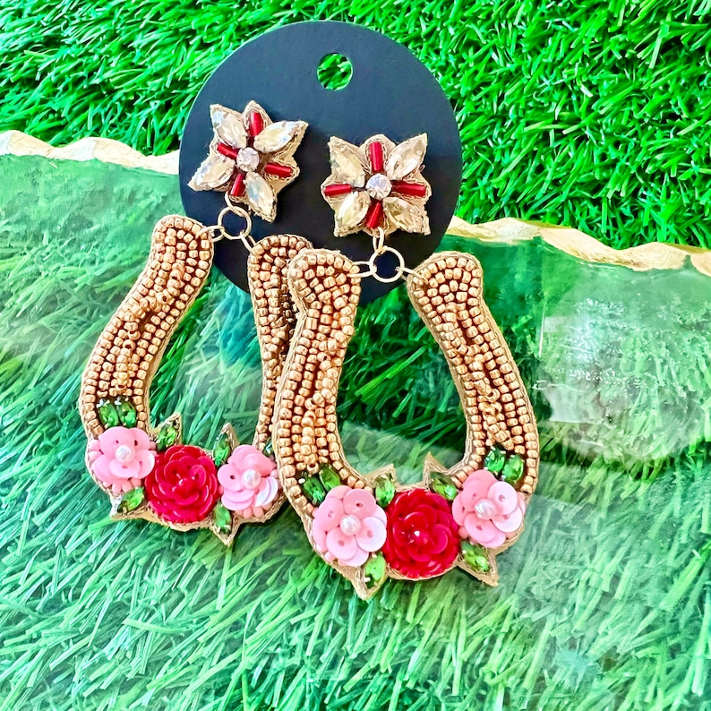 May include: A pair of gold horseshoe-shaped earrings with pink and red flower embellishments. The earrings are beaded and have a dangling flower charm.