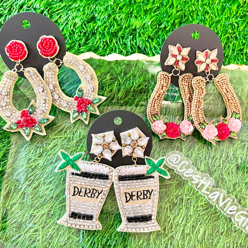 May include: Three pairs of beaded earrings, each with a different design. The first pair features a horseshoe shape with red and green beads. The second pair features a horseshoe shape with gold and pink beads. The third pair features a cocktail glass with black and white beads and the word "Derby" written on it.