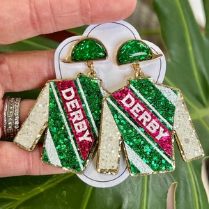 May include: A pair of green and white glitter earrings with a pink stripe and the word "Derby" on each earring. The earrings are shaped like a jockey's silks.