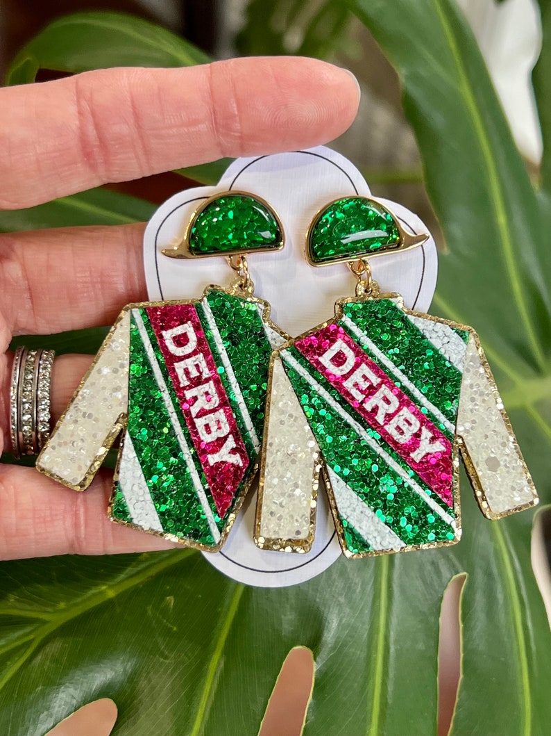 May include: A pair of green and white glitter earrings with a pink stripe and the word "Derby" on each earring. The earrings are shaped like a jockey's silks.