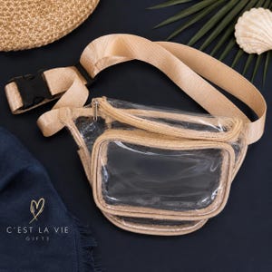 Puede incluir: Una riñonera de plástico transparente con bordes de tela beige, una correa ajustable a juego en color beige con una hebilla de plástico negra y dos compartimentos con cremallera. El frente tiene un bolsillo rectangular transparente.
