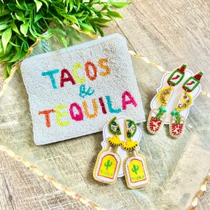 May include: A white beaded pouch with the text "Tacos & Tequila" in colorful beads. Two pairs of beaded earrings are displayed on a white card. One pair features a tequila bottle charm with a cactus and the text "Tequila" in gold beads. The other pair features a hot sauce bottle charm with a red chili pepper and a lime wedge.
