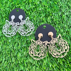 May include: A pair of gold and silver earrings featuring a tiger head design. The earrings are on a black background.