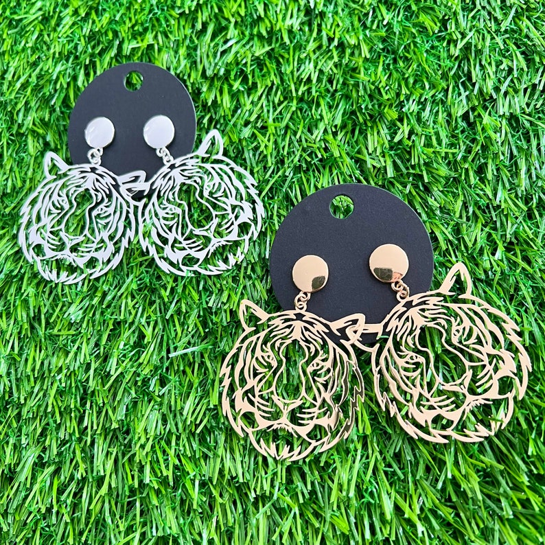 May include: A pair of gold and silver earrings featuring a tiger head design. The earrings are on a black background.