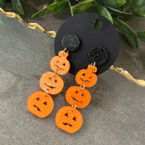 May include: A pair of dangle earrings featuring black glitter and orange pumpkin charms. Each earring has four pumpkin charms, each with a different jack-o'-lantern face.