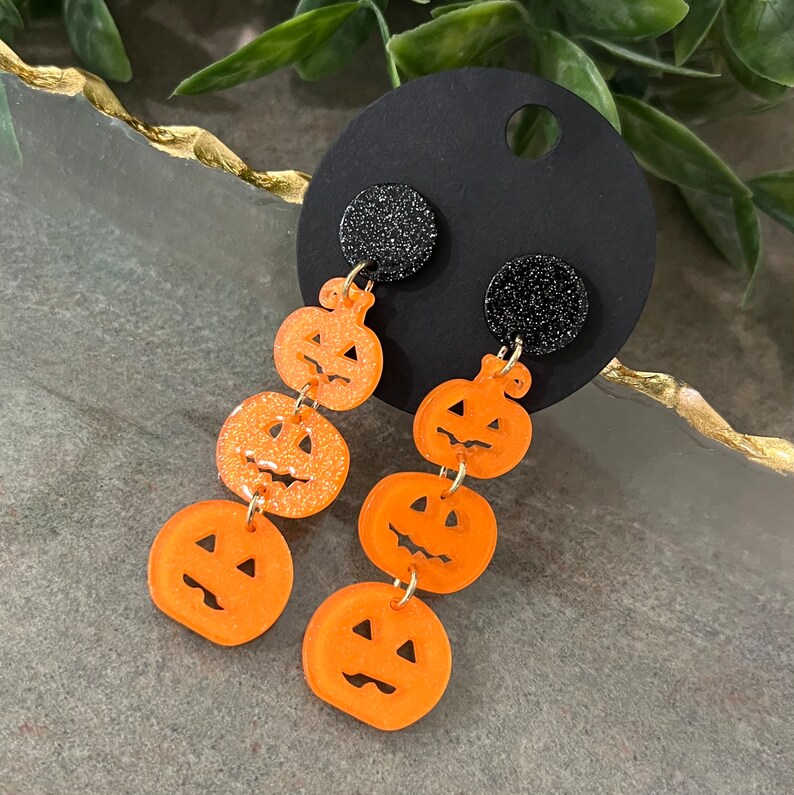 May include: A pair of dangle earrings featuring black glitter and orange pumpkin charms. Each earring has four pumpkin charms, each with a different jack-o'-lantern face.