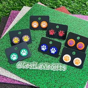 May include: A collection of seven pairs of circular stud earrings, each featuring a paw print design in various color combinations including orange, green, blue, purple, and red, displayed on black cards with the text @CestLaVieGifts visible.