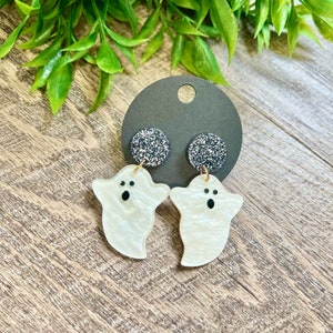 May include: A pair of white ghost-shaped earrings with black eyes and a sparkly black backing.