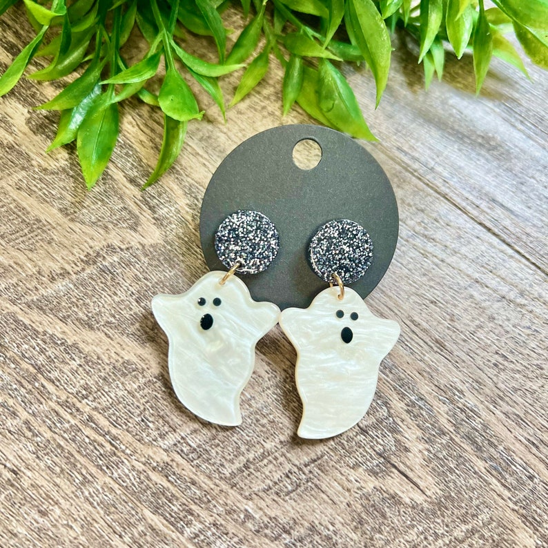 May include: A pair of white ghost-shaped earrings with black eyes and a sparkly black backing.