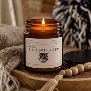 May include: A lit soy wax candle in an amber glass jar with a black lid. The white label features the text C'est la Vie Gifts, 100% natural, Smells Like A WILDCATS WIN, 8oz soy wax candle, 50 hours, and a graphic illustration of a wildcat head.