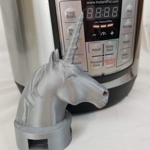 May include: A silver 3D printed unicorn head with a horn, sitting on a silver base with a rectangular opening. The unicorn head is positioned in front of an Instant Pot Lux Mini electric pressure cooker with a digital display showing 8:8:8.