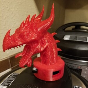 May include: A red 3D printed dragon head is used as a knob for a pressure cooker. The dragon has a textured surface and a detailed design. The pressure cooker is black with a silver rim.