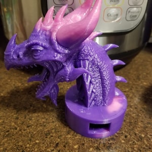 May include: A purple and pink 3D printed dragon figurine with a textured surface. The dragon has a long snout, sharp teeth, and large wings. The figurine is standing on a round base.