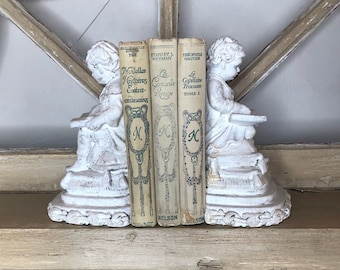 May include: White, distressed-finish bookends featuring cherubic figures, flanking three vintage books. The books have aged covers with French titles and the publisher's name, Nelson. The bookends and books sit on a wooden shelf.