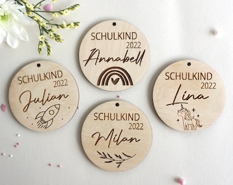 Pode incluir: Quatro ornamentos de madeira com o texto "Schulkind 2022" e um nome, cada um com um design diferente. Os designs incluem um foguete, um arco-íris, um unicórnio e um ramo.
