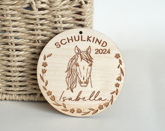 Pode incluir: Um ornamento redondo de madeira com uma ilustração de um cavalo, o texto "SCHULKIND 2024" e o nome "Isabella" gravados nele.