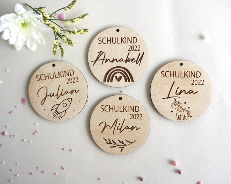 Puede incluir: Cuatro adornos de madera con el texto "Schulkind 2022" y un nombre, cada uno con un dise&ntilde;o diferente. Los dise&ntilde;os incluyen un cohete, un arco&iacute;ris, un unicornio y una rama.
