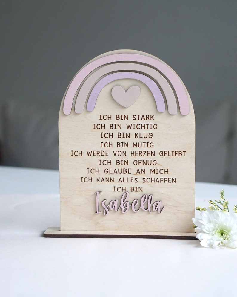 May include: A wooden rainbow with a pink heart in the center. The rainbow is painted in shades of pink and beige. The text on the sign reads: "ICH BIN STARK, ICH BIN WICHTIG, ICH BIN KLUG, ICH BIN MUTIG, ICH WERDE VON HERZEN GELIEBT, ICH BIN GENUG, ICH GLAUBE AN MICH, ICH KANN ALLES SCHAFFEN, ICH BIN Isabella"
