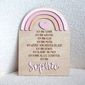 May include: A wooden sign with a pink and orange rainbow and a heart at the top. The sign has the text "ICH BIN STARK, ICH BIN WICHTIG, ICH BIN KLUG, ICH BIN MUTIG, ICH WERDE VON HERZEN GELIEBT, ICH BIN GENUG, ICH GLAUBE AN MICH, ICH KANN ALLES SCHAFFEN, ICH BIN Sophie" in German.