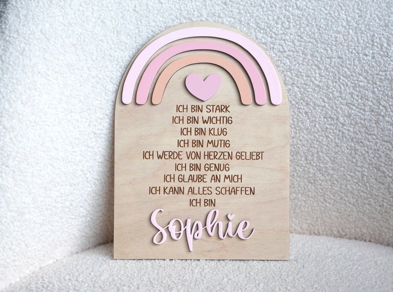May include: A wooden sign with a pink and orange rainbow and a heart at the top. The sign has the text "ICH BIN STARK, ICH BIN WICHTIG, ICH BIN KLUG, ICH BIN MUTIG, ICH WERDE VON HERZEN GELIEBT, ICH BIN GENUG, ICH GLAUBE AN MICH, ICH KANN ALLES SCHAFFEN, ICH BIN Sophie" in German.
